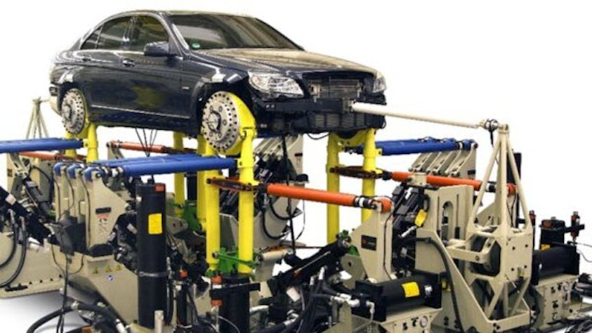 A vehicle is mounted on a large, complex automotive testing rig with yellow supports, blue beams and various hydraulic components, suggesting vehicle performance or durability testing.