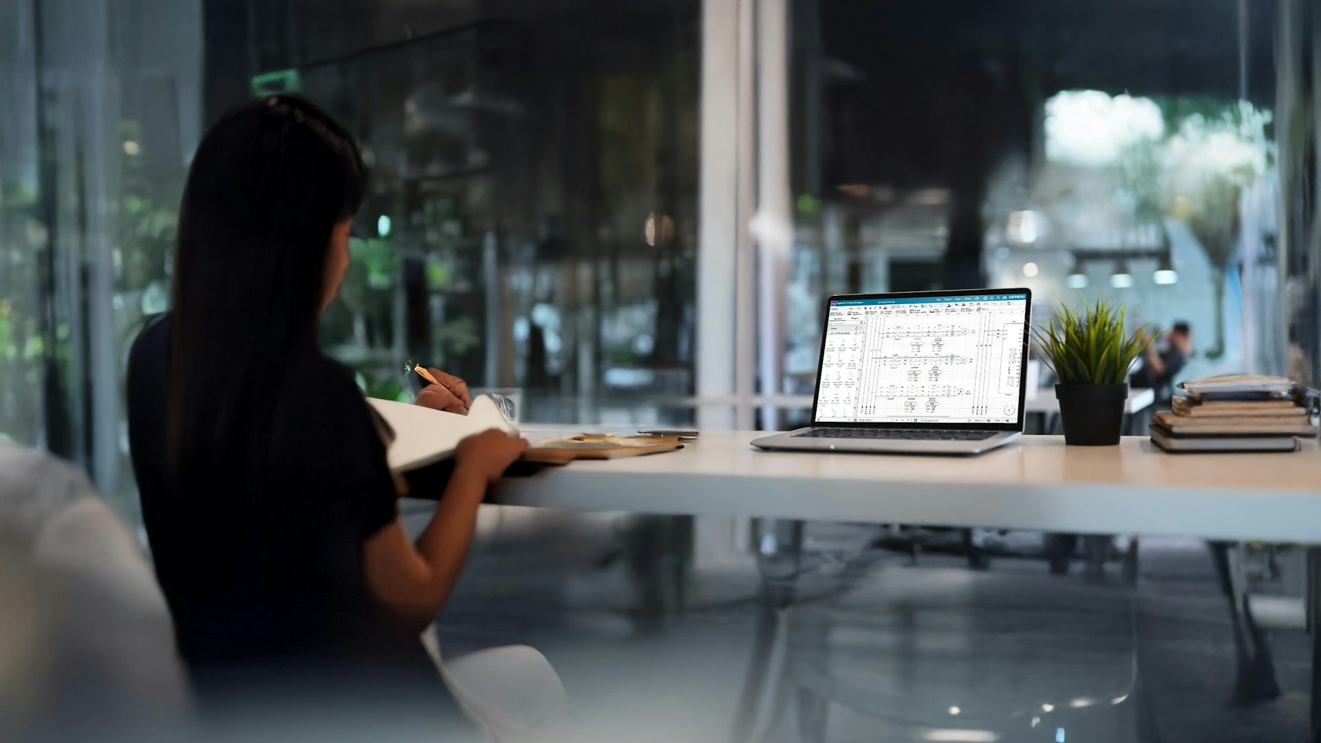 Woman at desk writing while viewing a electrical diagram on her laptop.