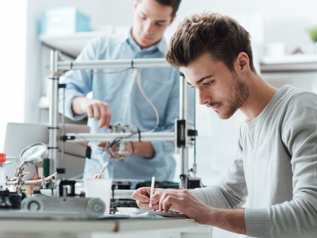 Two men in a robotics lab, one is working on a machine and the other is taking notes Two men in a robotics lab, one is working on a machine and the other is taking notes