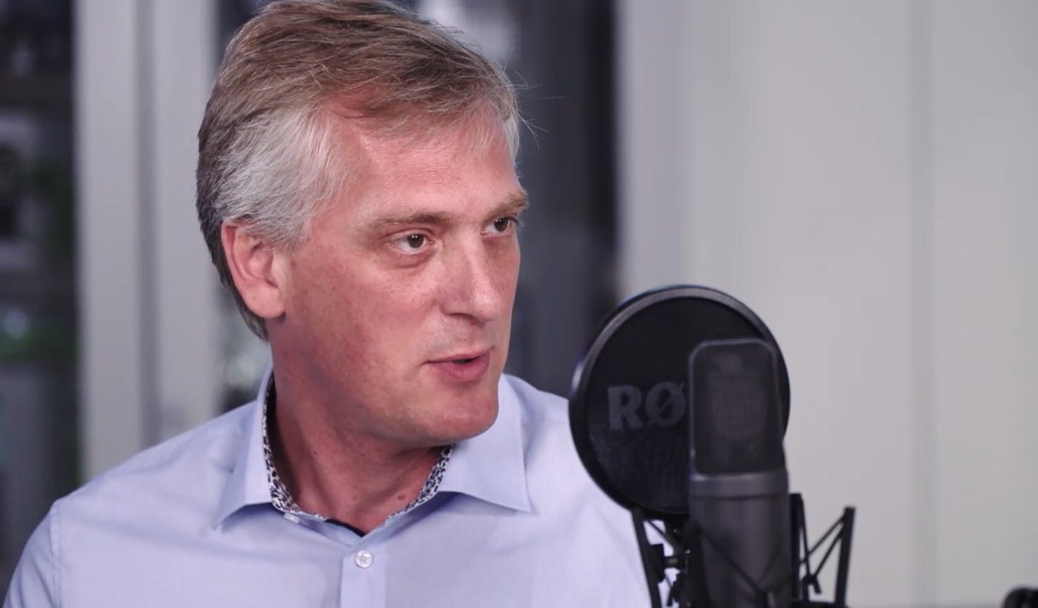 Professional speaker recording a podcast episode, wearing a light blue business shirt in front of a studio microphone