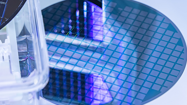Close-up of a silicon wafer with blue and purple illumination showing grid pattern of squares. A transparent container or testing apparatus is visible in the foreground.