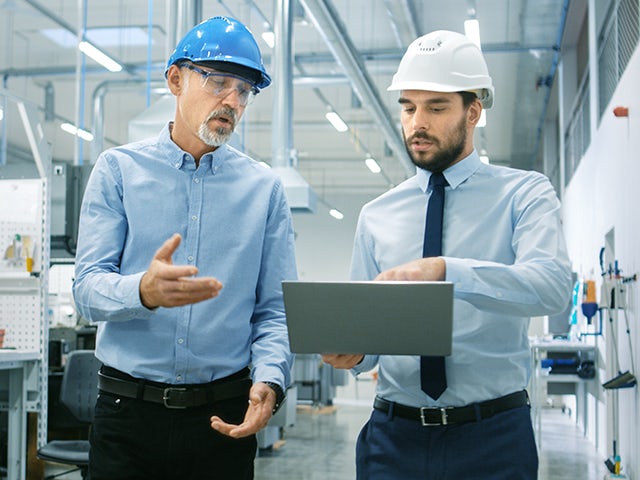 An engineer shares project information on a laptop with another engineer while they walk through a modern factory. The project information includes a CAD model with information the manufacturing process for the part they are displaying.