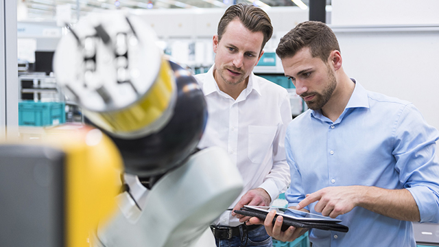 Two people inspect a factory robot arm, with one of the people holding a tablet computer