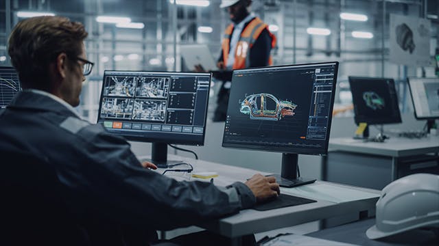 Operator working at desk with two desktop monitors, overviewing car production in a car assembly plant.