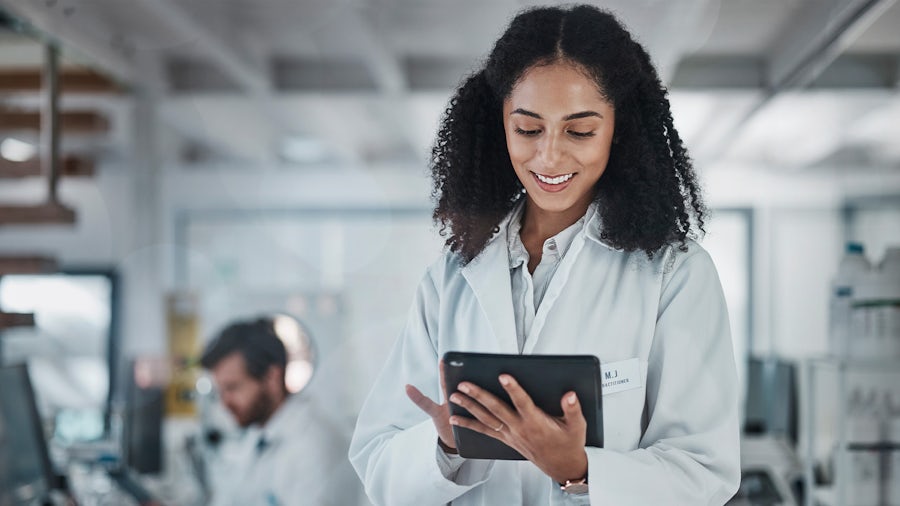 A female medical professional wearing a white lab coat is holding and looking at a tablet