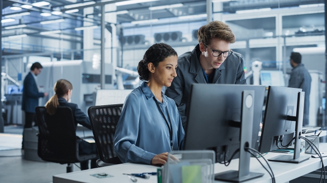 Engineers collaborating on a project using a computer in a factory with robotic arms producing devices for the tech industry.