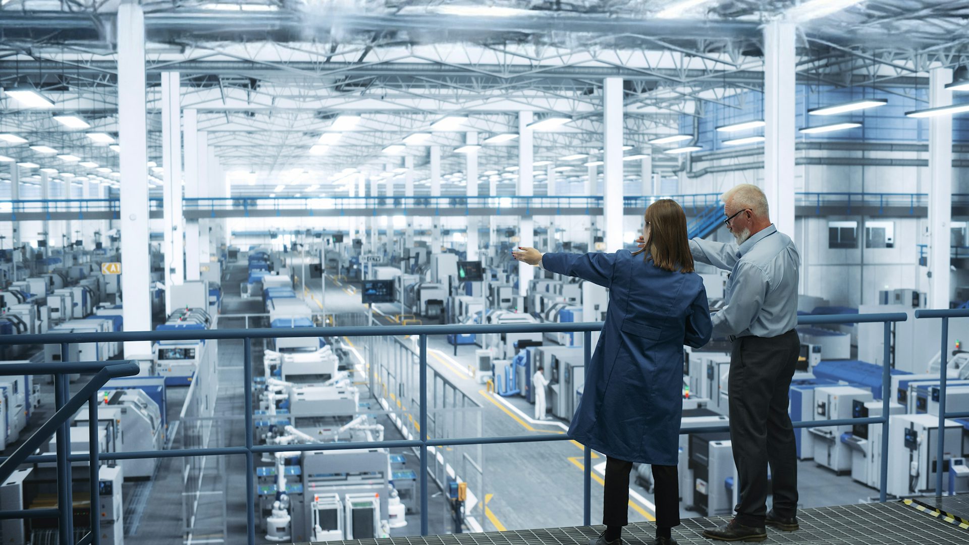 Male and female engineers standing on a platform with their back to camera, using a laptop and discussing production at a modern AI automated electronics plant.