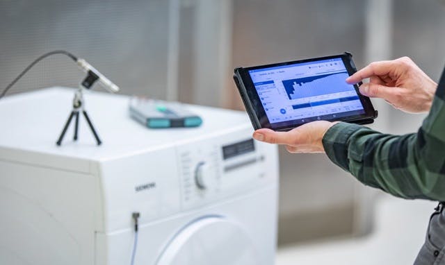 Engineer performing sound and vibration testing on a washing machine, viewing the results on a tablet.