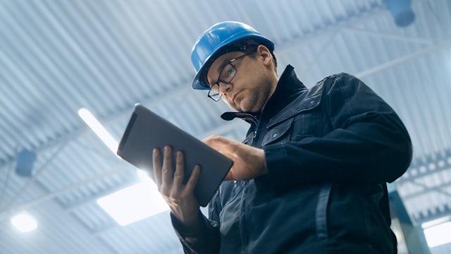 A man wearing a hard hat, glasses and a jacket while looking at an iPad, checking product documentation.