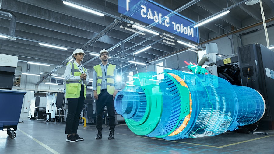 Two engineers wearing safety vests and hard hats stand in a smart factory, analyzing a large digital model of machinery using augmented reality software.