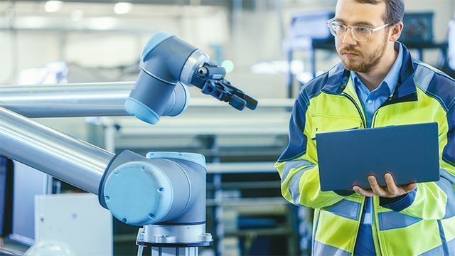 Hombre que inspecciona un robot de fábrica mientras sostiene un ordenador portátil.