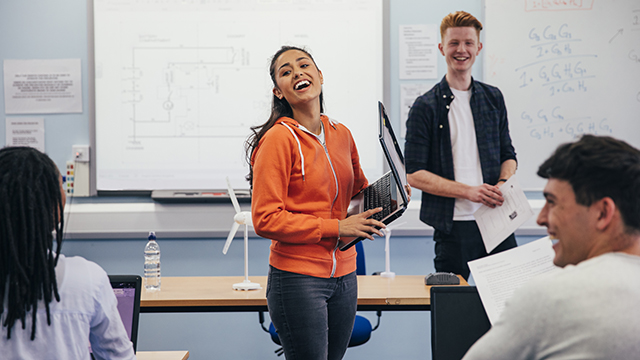 한 젊은 여성이 노트북을 들고 학생들에게 자신의 작품을 발표할 준비를 하며 웃고 있습니다. 그녀는 주황색 집업 후드티를 입고 있으며, 그녀와 이야기하는 이미지의 배경에는 또 다른 남학생이 있습니다. 두 명의 학생이 책상에 앉아 프레젠테이션을 보고 있는 모습이 이미지의 맨 앞에 나와 있습니다.