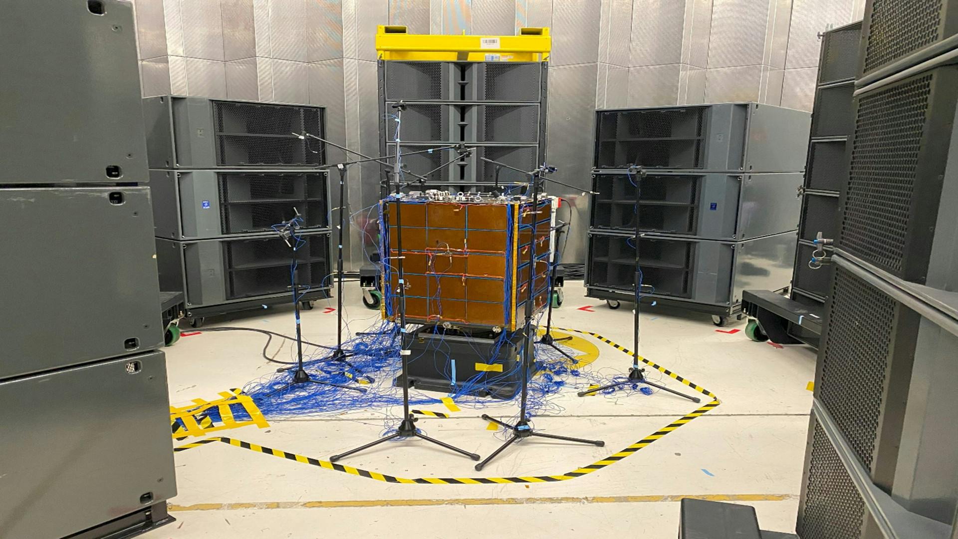 Spacecraft surrounded by speakers and sensors during Direct Field Acoustic Noise (DFAN) testing setup.