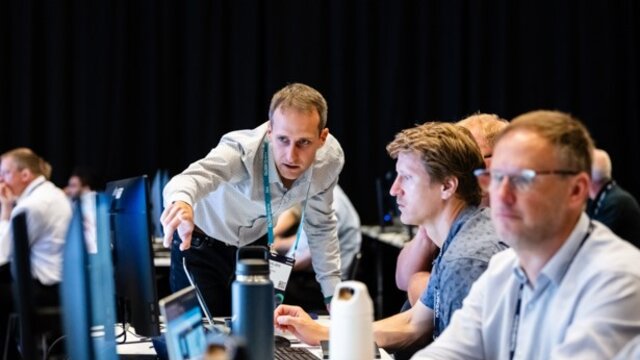 Several men sitting at computers during a hands-on-training session at Realize LIVE Europe with the presenter assisting them.