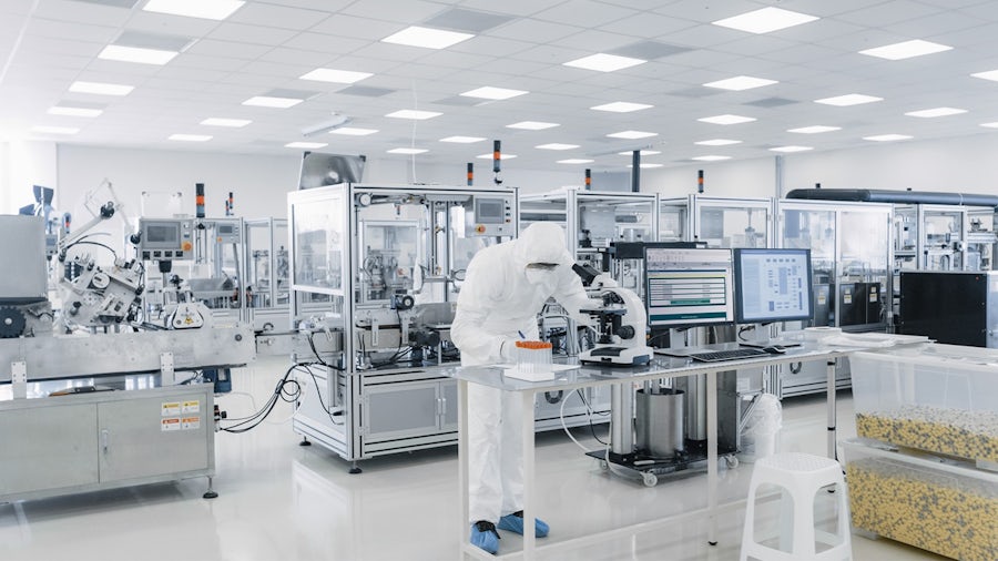 Scientist in cleanroom suit operating a microscope and computer in a bright, sterile pharmaceutical manufacturing plant with automated machinery.