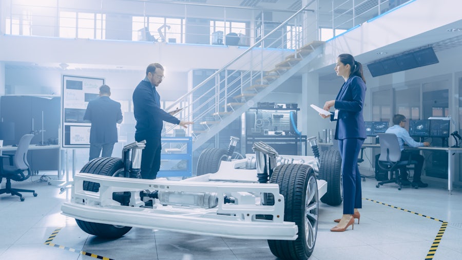 A man and woman discuss components of a car chassis on the floor between them.