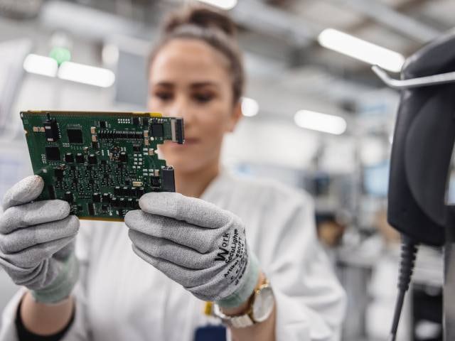 Une personne portant des gants tient un circuit imprimé devant la caméra. Le circuit imprimé semble être destiné à un type d'appareil électronique ou informatique.