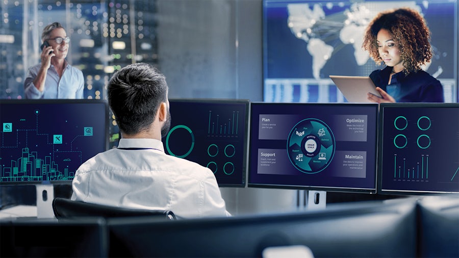 Man in white shirt monitors a multi-screen display with other workers talk on the phone and work on a tablet