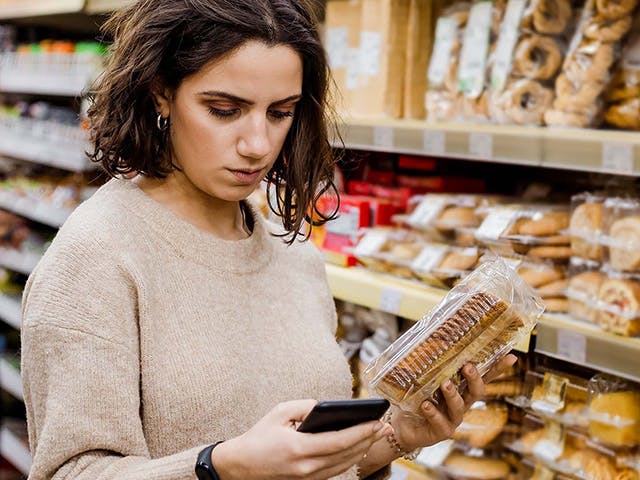 Frau mit einer Schachtel Cracker im Gang eines Lebensmittelgeschäfts schaut auf ihr Handy.