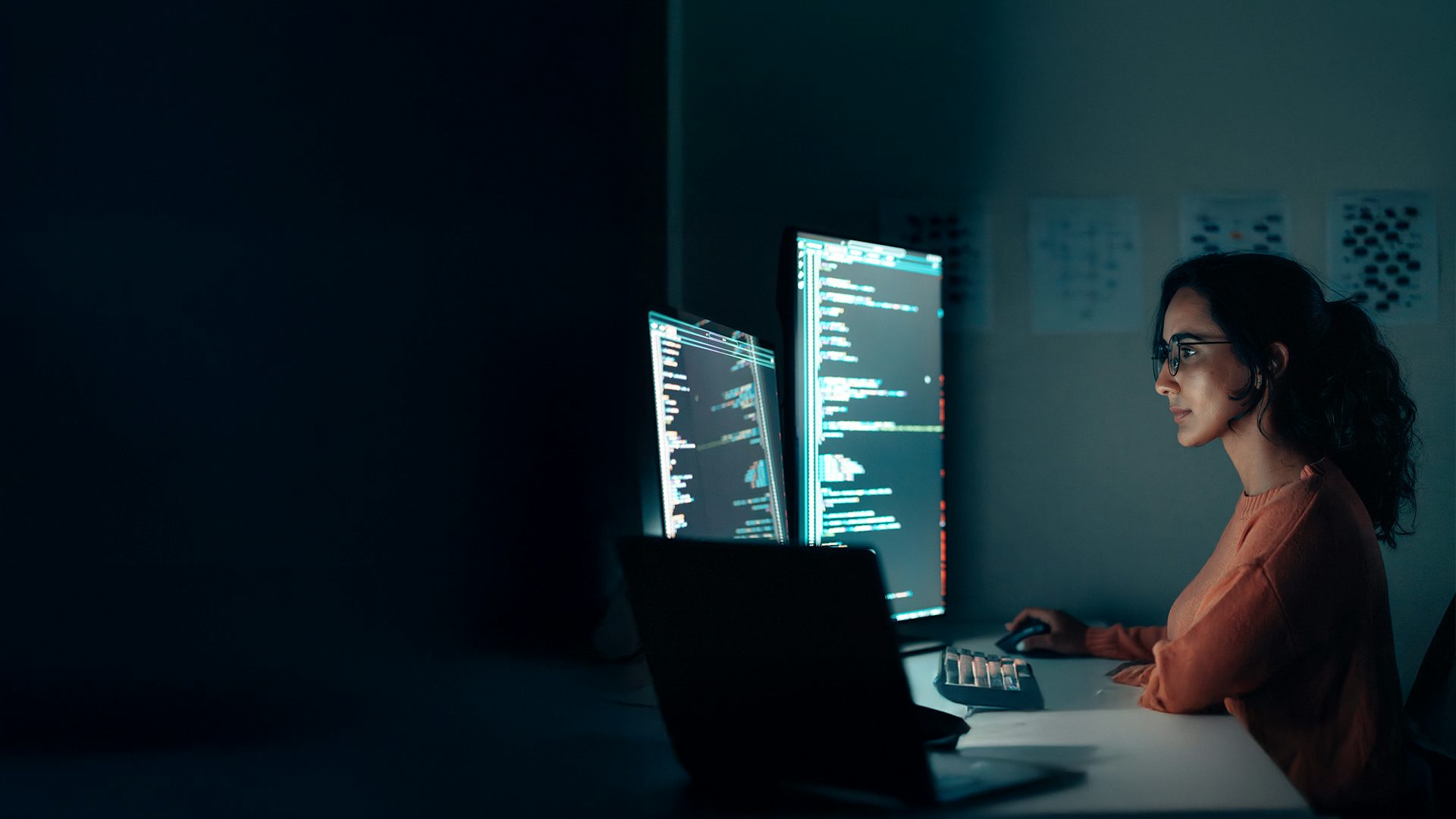 A female programmer coding at night in a high-tech workspace with a laptop and multiple monitors.