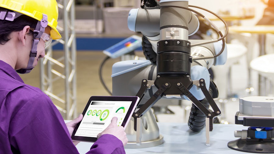 Smart manufacturing equipment and robotics in an industrial setting. A person in a purple uniform is operating a tablet connected to a robotic arm, suggesting advanced automation and digital control in the manufacturing process.