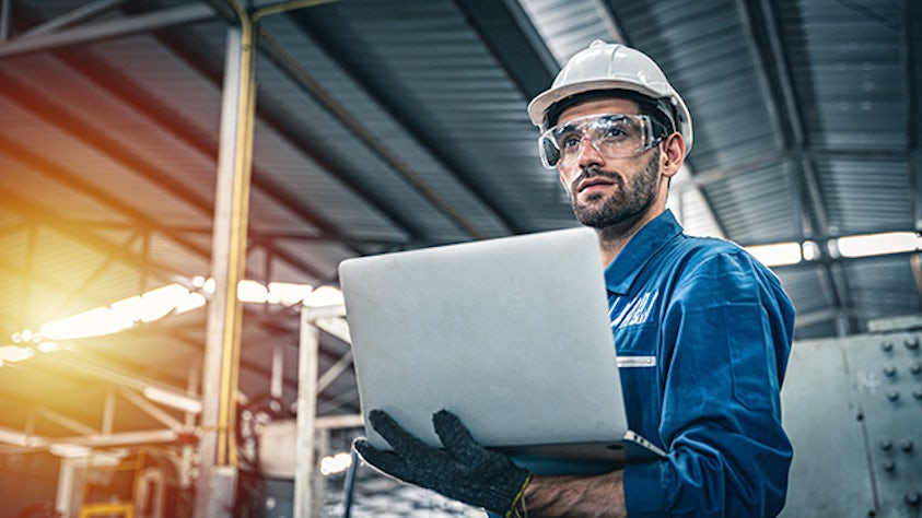 A man with a hardhat in a factory holds a laptop.
