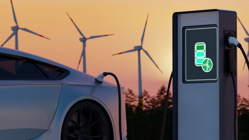 A silver electric vehicle parked at a charging station, while three windmills turn at sunset.