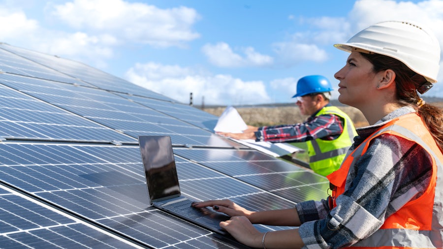 An engineer with a laptop sat on a solar panel observing the solar panel. 