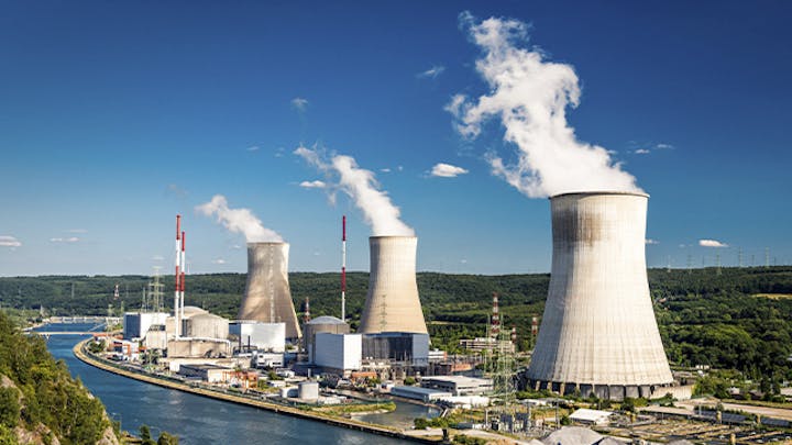 Aerial view of a nuclear power plant complex with multiple cooling towers emitting steam, surrounded by a forested landscape and a river or body of water.