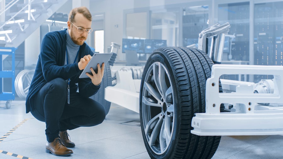 An engineer on the shop floor examining a tablet while crouching next to the right, front tire on a vehicle frame. 
