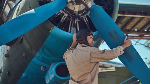 A pilot mechanic in full flight gear checks the propeller on a retro military aircraft