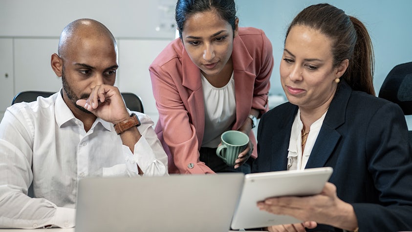 Image of 3 co-workers looking at a computer monitor and a laptob in an office setting. This represents the concept of APQP quality project management.
