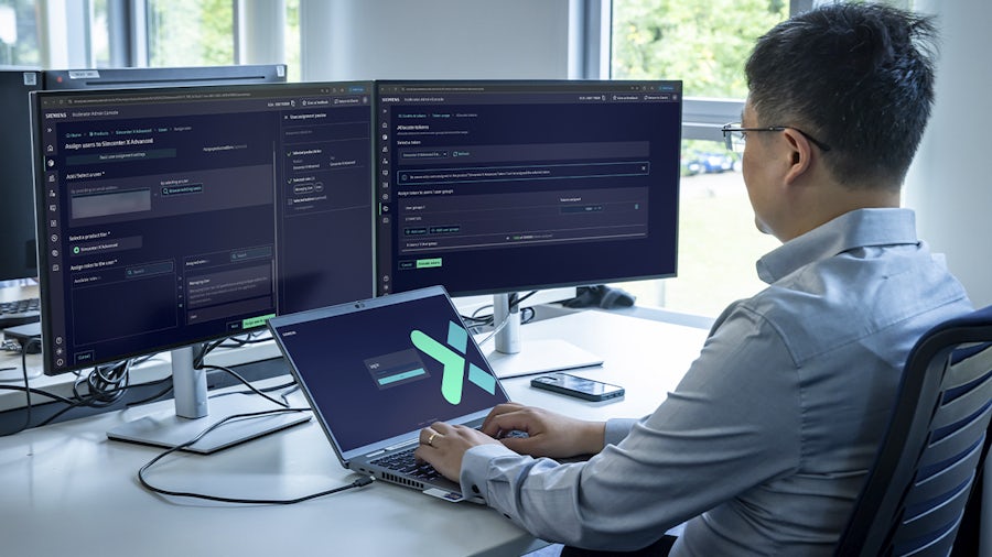 An engineer using a laptop and two monitors to manage user assignments and token allocation within the Siemens Xcelerator Admin Console.