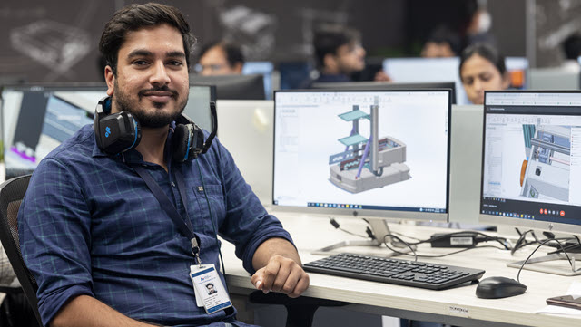 Engineer is looking to the camera and smiling while sitting on a desk with two monitors