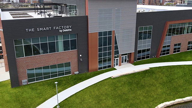 Exterior view of The Smart Factory by Deloitte, a modern brick and glass building with large windows, a rooftop terrace, and a curved sidewalk leading through a green lawn.
