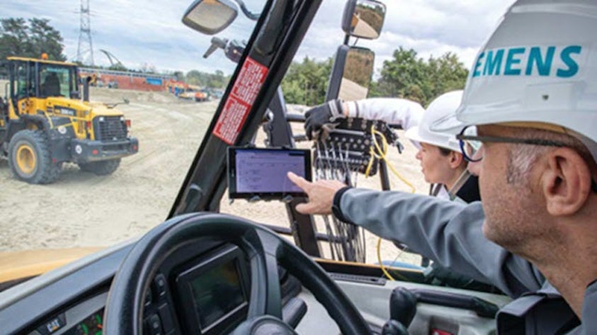 An engineer wearing a Siemens hard hat points at a tablet displaying data inside a heavy machinery cab, with another loader visible at a dusty test site in the background.