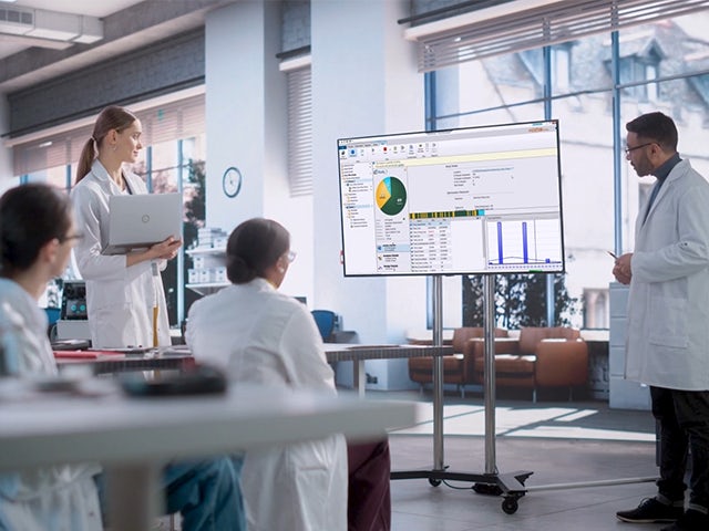 A group of laboratory professionals in white lab coats observing a presentation displayed on a large screen.