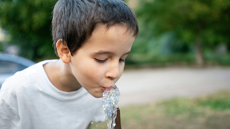 A young boy with dark hair and a white t-shirt drinks from a water fountain, eyes closed, as water splashes into his mouth. The background is blurred green foliage and light ground.