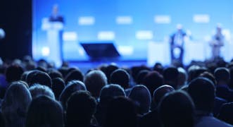 Abbildung eines großen Publikums, das einem Redner auf einer Bühne zusieht.