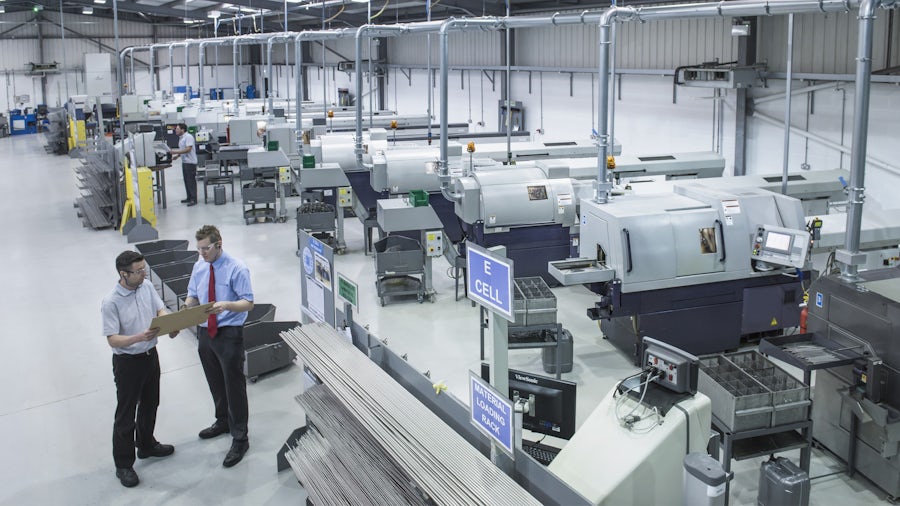 Two engineers stand on the factory floor looking at manufacturing plans.