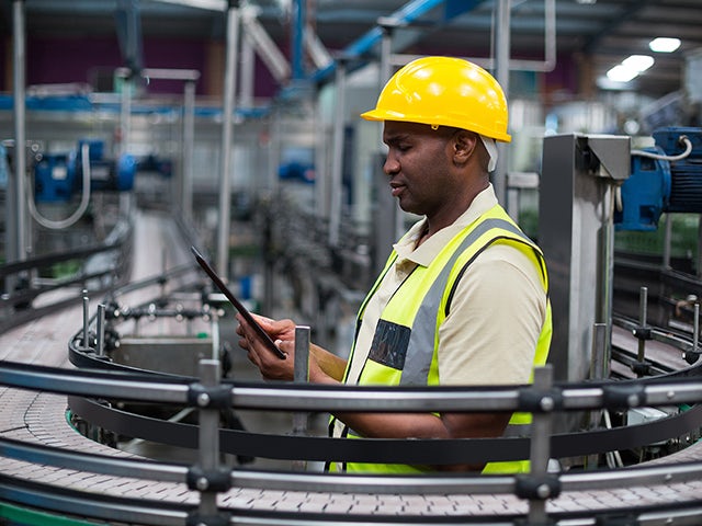 Quality management professional in a yellow hard hat and reflective vest uses a digital tablet to oversee an industrial production line.