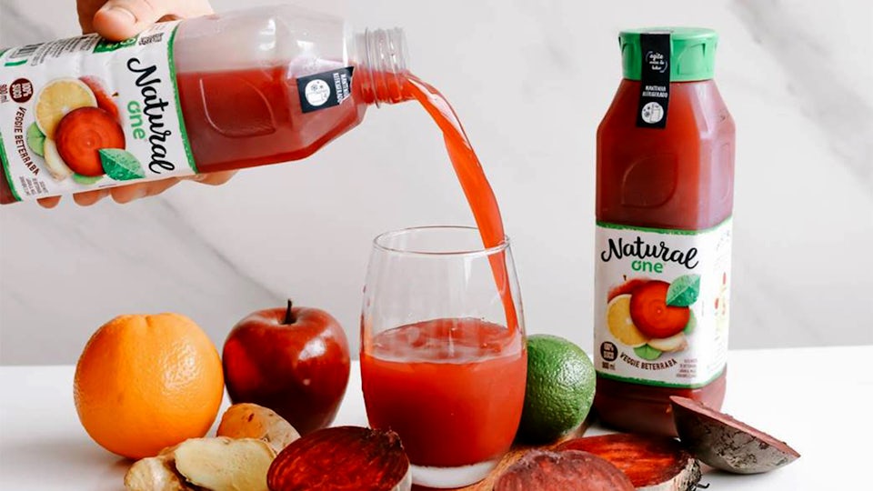 A bottle of Natural One juice is poured into a glass, next to various fruits and another unopened bottle of juice