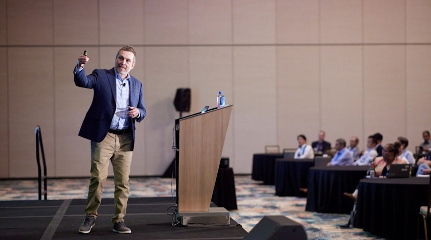 Speaker standing on stage pointing to a screen at a user event.