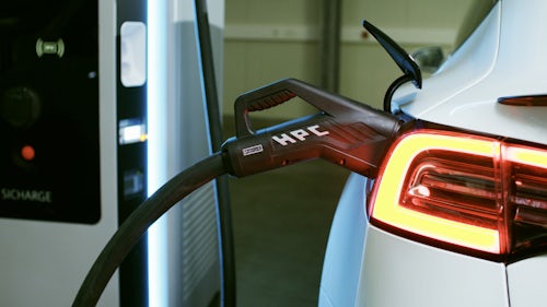 Close up photo of a charging cable plugged into the port of a white electric car with an illuminated brake light. 