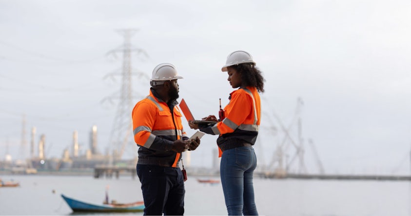 Two engineers in hard hats and safety vests with a laptop at a construction site.