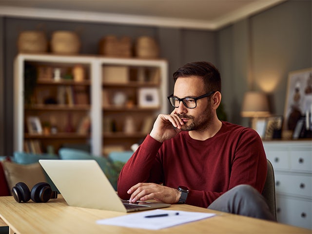 A young adult male sitting at a desk and resting his head on his hand while working on a project on his laptop.