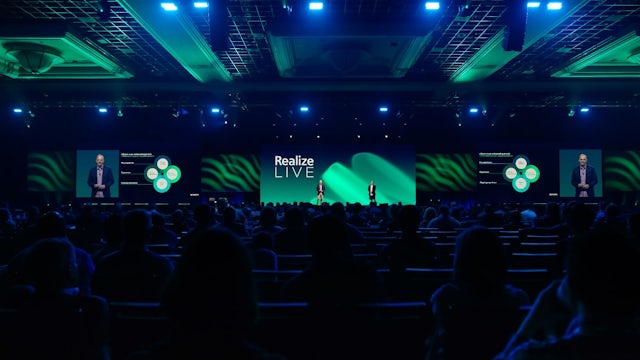 The main general session with 2 people on stage and people sat down in the audience at the digital transformation conference, Realize LIVE