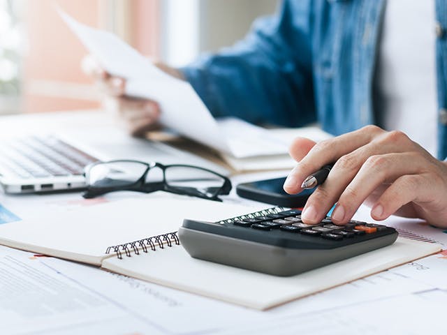 A person sitting in front of a desk holding a document on the left hand and a calculator on the right one.