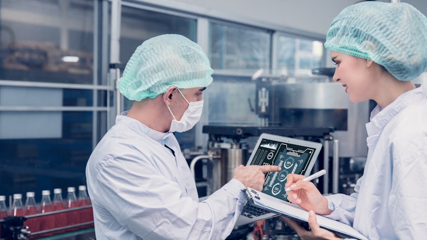 Man and woman looking at a tablet in a food & beverage factory wearing a hair net and a white lab coat.