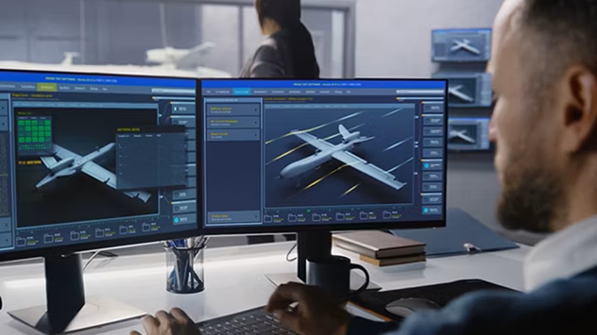 A man is working on a dual-monitor setup displaying 3D models and system data for an aircraft in a high-tech office environment.